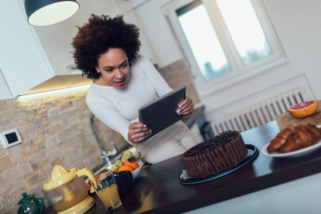 Woman uses iPad camera to take detailed photo of a chocolate cake on kitchen counter.