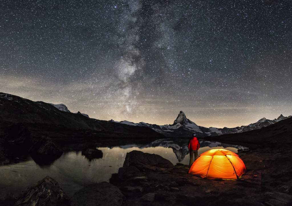 A photo of person in red next to well-lit tent under Milky Way is taken with iPhone Night mode.