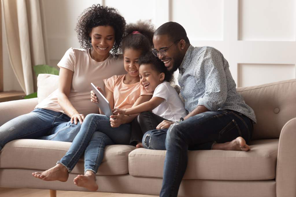 An African American family gathers on couch with iPad, smiling about their Apple photo book.
