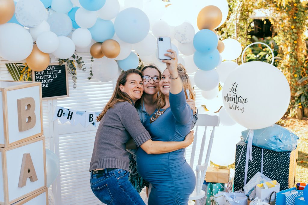 Female friends taking selfie with pregnant woman at a baby shower. Mobile photography, party decorations in white and blue colors, baby boy